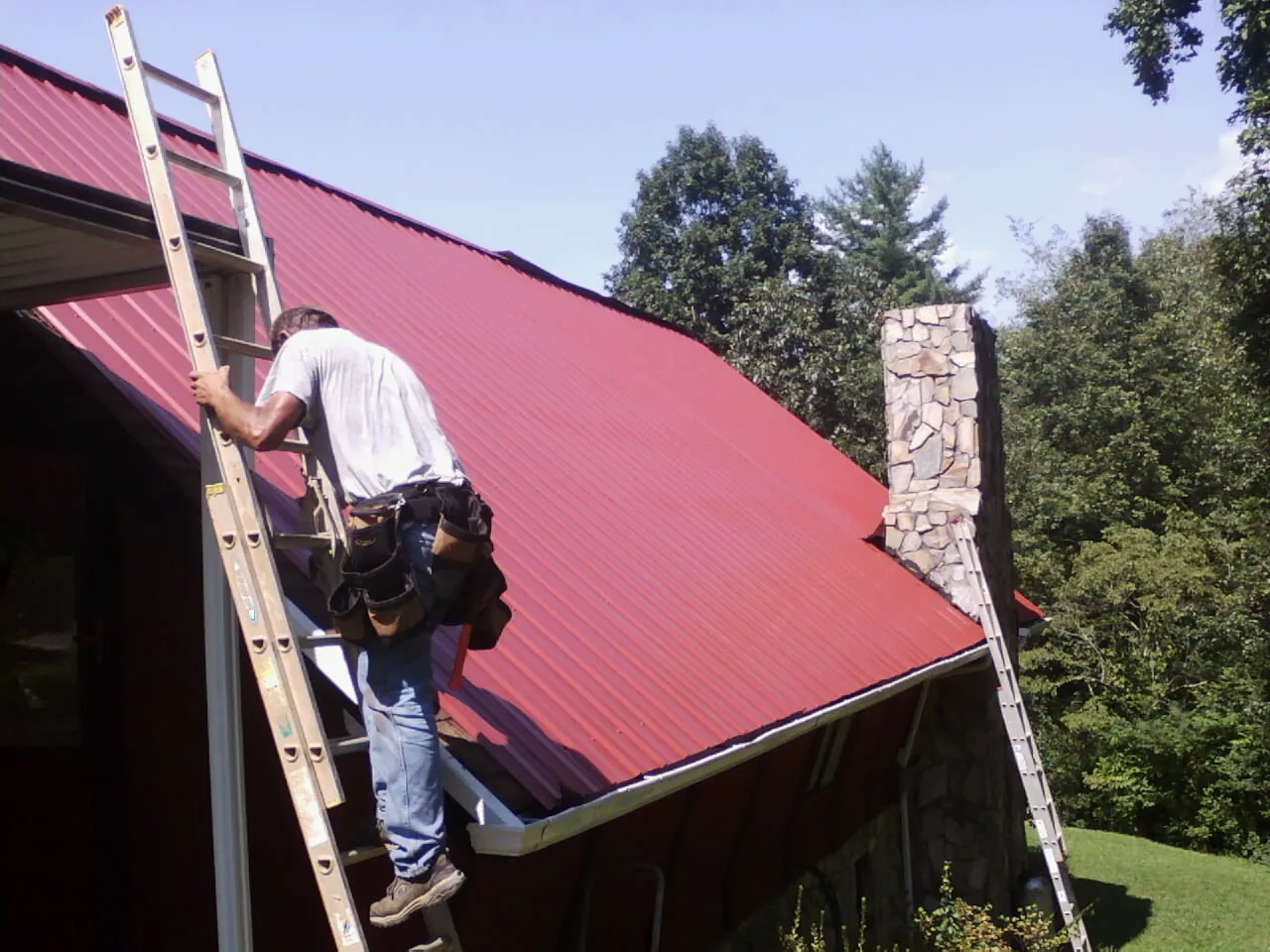 Roofer on a ladder beside a red metal roof and stone chimney — Soffit & Fascia Repair by Roofing Brothers in Thompsontown